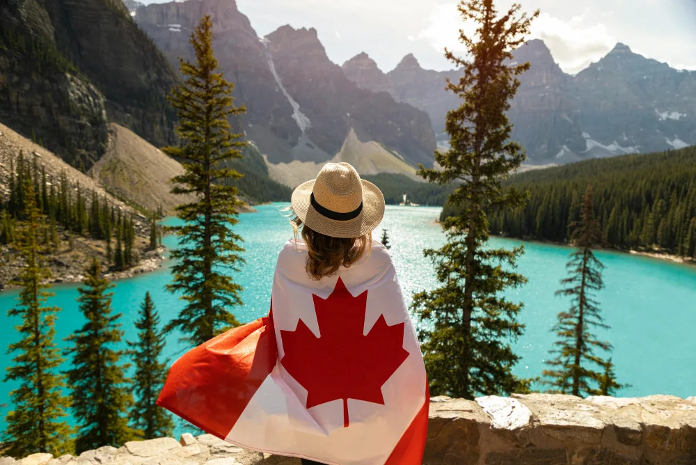 A traveller in a sun hat holding a Canadian flag above a turquoise glacial lake ringed by pines